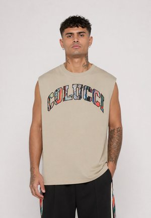 Mann med tatoveringer har på seg en ermeløs beige skjorte med fargerik "Colucci"-tekst, kombinert med svarte bukser med mønstrede striper.