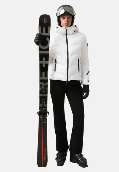 Witte geïsoleerde skijack met hoge kraag, zwarte skibroek, zwarte handschoenen en ski met een rode accent. Draagt een zwarte helm en skibril.