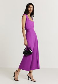 Jumpsuit senza maniche di colore viola con pantaloni a gamba larga, texture liscia, vita stretta. La modella tiene una piccola borsa a tracolla nera in vernice, indossando tacchi a strappo neri.
