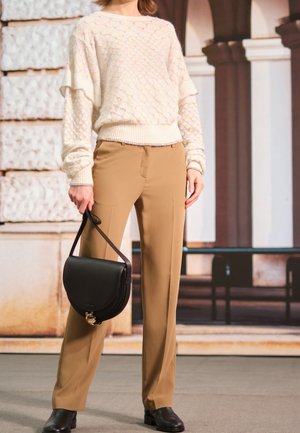 Persona con un suéter crema texturizado y pantalones anchos color tostado, sosteniendo un bolso de mano de cuero negro, de pie en el interior junto a una pared de piedra.
