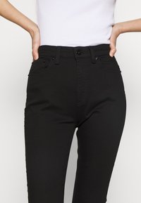 Mujer que lleva pantalones negros de talle alto y una blusa blanca ajustada sin mangas, con las manos en las caderas.