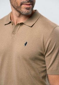 Férfi bézs rövid ujjú galléros pólóban, két gombbal és a mellkason egy kis sötét emblémával, arcszőrzettel és ajkakkal látható.