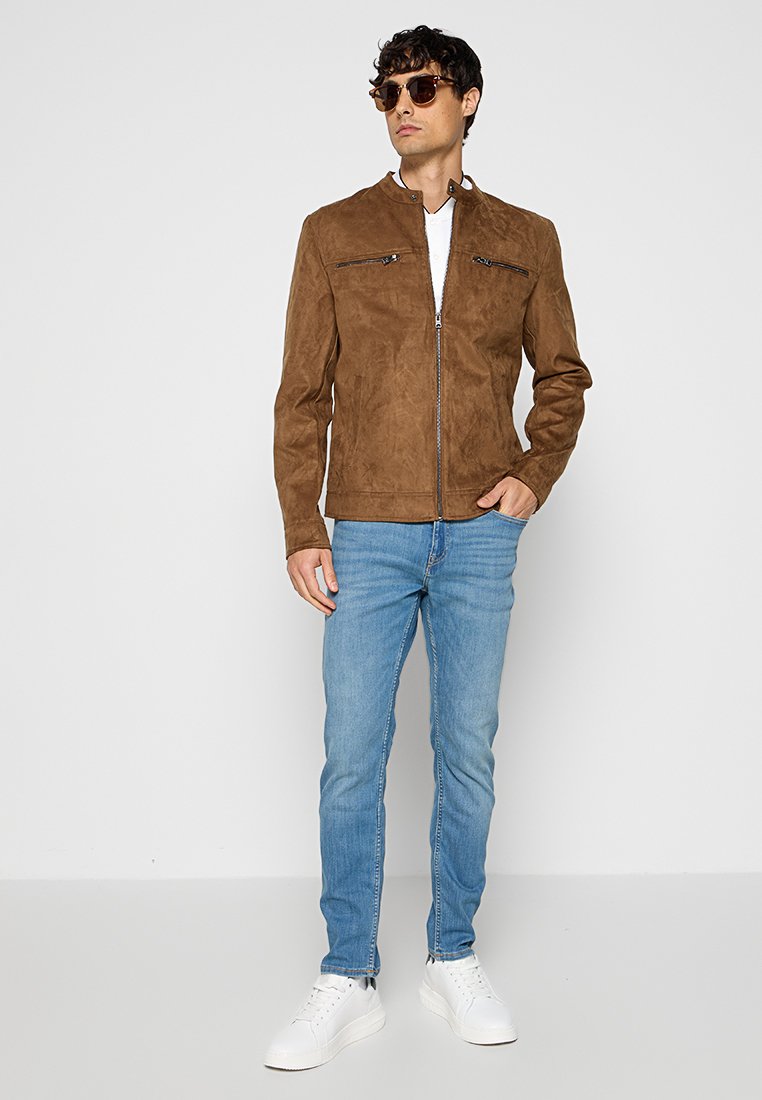 Veste en daim marron avec fermeture éclair à l'avant, deux poches zippées poitrine, col rond, associée à un jean bleu clair et des baskets blanches.