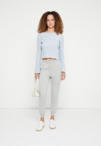 Lichtblauw geribd top met lange mouwen en een cropped zoom, gecombineerd met grijze trainingsbroek en witte sneakers. Model houdt een kleine beige handtas vast.