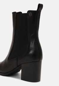 Černá kožená kotníková bota s elastickým bočním panelem, poutkem, kulatým stříbrným emblémem značky a blokovým podpatkem na bílém pozadí.