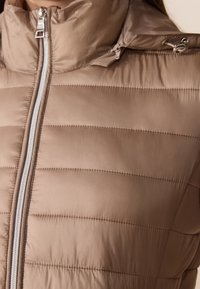Beżowa pikowana kurtka z wysokim kołnierzem, pionowym pikowaniem oraz srebrnym zamkiem błyskawicznym. Posiada boczną kieszeń i gładką, elegancką teksturę.