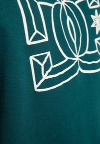 Tejido de algodón en color verde azulado con bordados geométricos en blanco que presentan motivos de hojas y estrellas, destacando una superficie texturizada y elementos de diseño atrevidos.