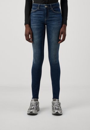 Persoon die donkerblauwe skinny jeans en grijze sportsneakers draagt, staand tegen een effen lichte achtergrond.