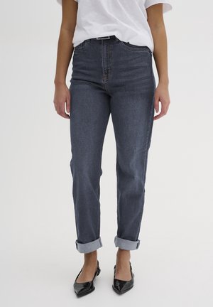 Mujer con vaqueros azul oscuro con puños, camiseta blanca y zapatos planos negros de punta puntiaguda, de pie frente a un fondo liso.