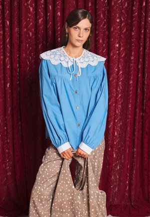 Jeune femme en blouse bleue à col en dentelle blanche et pantalon beige à pois, tenant un sac beige assorti devant des rideaux en dentelle rouge.