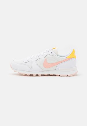 Biały sneaker Nike z różowym logo i żółtą wstawką na pięcie, z teksturowaną podeszwą i sznurówkami, widoczny z profilu.