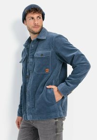 Blå fløjlskjorte med knaplukning, brystlommer og en struktureret overflade. Båret med grå jeans og en matchende hue.