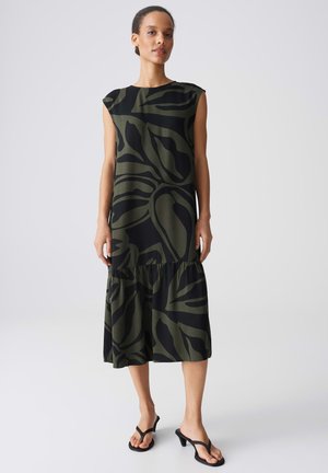 Frau steht und trägt ein ärmelloses, dunkelgrün-schwarz gemustertes Midikleid mit Volantsaum und schwarze Sandalen mit Absatz und Zehensteg.