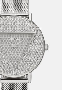 Reloj Guess plateado con correa de malla, caja redonda y una esfera triangular adornada con numerosos cristales transparentes y diales minimalistas.