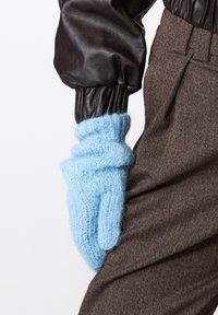 Šviesiai mėlyna nertinė pirštinė su pūkuota tekstūra, su laisvų kirpimų dizainu, dėvima ant rudos džinsinio audinio kelnių su aukštu juosmeniu.