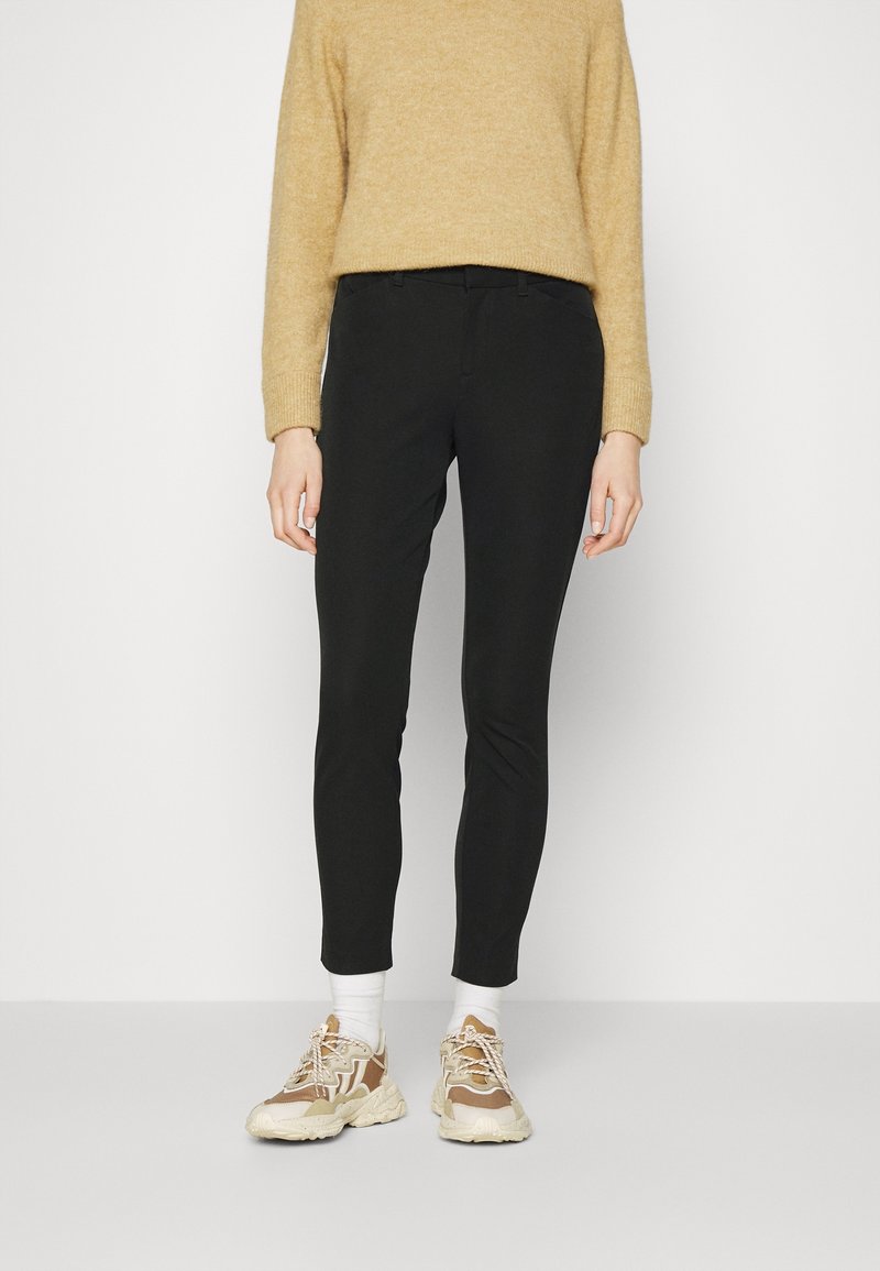 Pantalon noir slim en tissu doux, associé à un pull en maille beige et à des baskets de couleur claire avec des accents beige et crème.