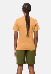 Osoba o długich, splecionych włosach, ubrana w beżową koszulkę z krótkim rękawem i oliwkowo-zielone spodenki, stojąca tyłem na jednolitym tle.