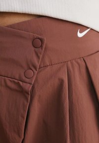 Bruna plisserade shorts i en texturerad tyg, med två bruna knappar över midjan och en vit Nike-logotyp på övre kanten.