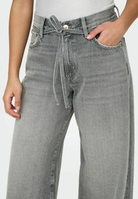Jeans larghi grigi con vita alta, dettaglio legato sul davanti e tasche laterali. Texture di denim sbiadito con cuciture a contrasto.