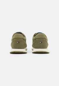 Pár olivově zelených textilních tenisek s bílými podrážkami a malým červeno-bílo-modrým logem na patě, zobrazeno ze zadní strany.