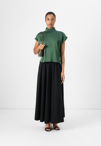 Haut vert à col montant et à manches courtes associé à une jupe longue et fluide noire. Le mannequin tient un petit sac à main marron. Design simple et minimaliste.