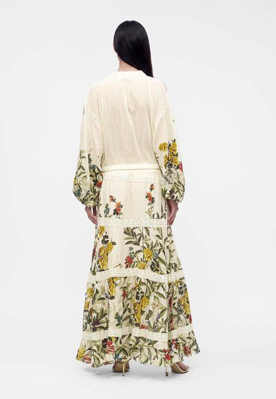 Femme aux longs cheveux foncés porte une longue robe fleurie crème avec manches longues et jupe à volants, debout de dos sur fond blanc.