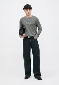 Joven con cabello rizado que lleva un suéter de punto gris, jeans anchos negros, cinturón negro y zapatos negros, sosteniendo un bolso de mano de cuero negro.