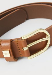 Ceinture en cuir marron avec une boucle en métal doré et un accent de logo. Texture lisse avec des coutures précises le long des bords.