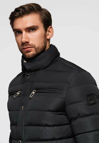 Černá péřová bunda s vysokým límcem, vybavená kapsami na zip na hrudi a kovovými akcenty. Prošívaná textura s elegantním designem.