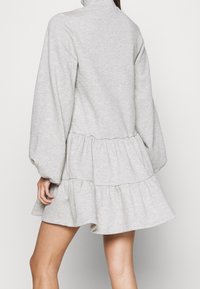 Femme portant une robe courte légère grise à manches longues et à volants, avec un col haut, vue de dos sur un fond uni.