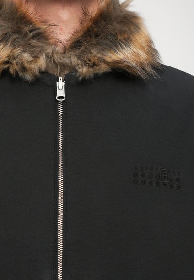 Svart jacka med pälsfodrad krage, frontdragkedja och subtilt texturerad logotype på bröstet.