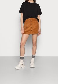 Mini falda de pana marrón con botones en la parte delantera, combinada con una camiseta negra de corte holgado y zapatillas. Tejido texturizado, diseño de talle alto.