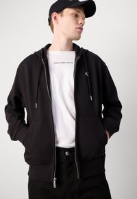 Sudadera negra con cremallera hecha de un tejido suave, que cuenta con capucha con cordón y bolsillos frontales. Lleva puesta una camiseta blanca de Calvin Klein Jeans.