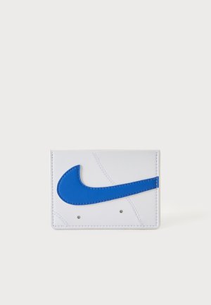 Baltas piniginė su dideliu mėlynu Nike swoosh logotipu, pritvirtintu priekinėje pusėje, rodomas ant vientisos šviesios fono.