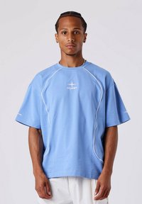 Camiseta de gran tamaño en azul claro de tejido suave, con acentos en blanco y un logo central. Las mangas cortas y el corte relajado enfatizan la comodidad.
