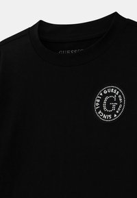 T-shirt nera in cotone con colletto rotondo. Presenta una toppa rotonda con il logo 'Guess' in bianco e decorazioni argentate.