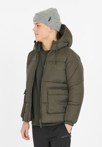 Olivegrøn pufferjakke med hætte, lynlås og forlommer. Struktureret stof med en glat finish. Båret over en mørk sweatshirt.