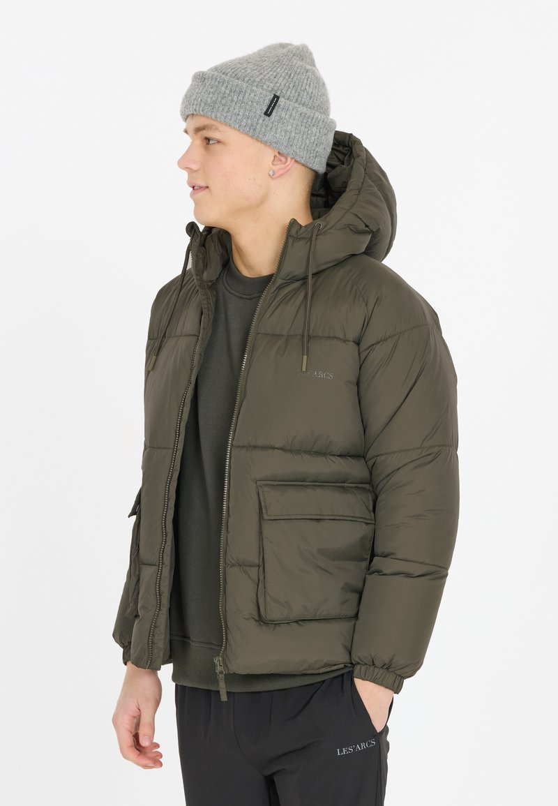 Olivegrøn pufferjakke med hætte, lynlås og forlommer. Struktureret stof med en glat finish. Båret over en mørk sweatshirt.