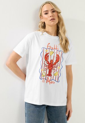 Blonde Frau trägt ein weißes T-Shirt mit rotem Hummer, Zitronenspalten und dem Schriftzug "Frutti di Mare", kombiniert mit blauen Jeans.