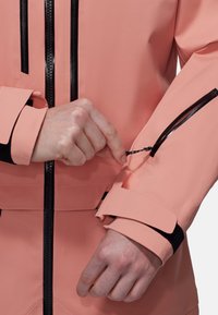 Helle rosa wasserfeste Jacke mit schwarzem Reißverschluss und Akzenten. Verfügt über verstellbare Klettverschlüsse an den Ärmelabschlüssen und eine Reißverschlusstasche am Arm.