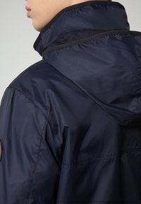 Giacca impermeabile blu navy con colletto alto, fodera in rete e cappuccio. Presenta una toppa rotonda con logo sulla parte superiore della manica. Texture liscia.