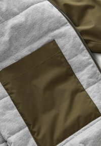 Szara kurtka puchowa z olivową, błyszczącą kieszenią. Posiada poziomy wzór pikowania i zamek błyskawiczny po boku.