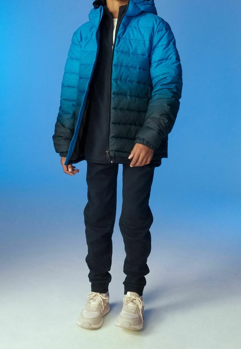 Doudoune bleue et noire avec capuche, motif texturé, portée sur une chemise sombre et associée à un jogging sombre et des baskets claires.