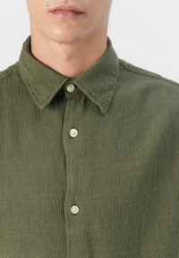 Olive green button-up shirt with a textured fabric, a pointed collar, and light-colored buttons. Features a relaxed fit and subtle pattern.