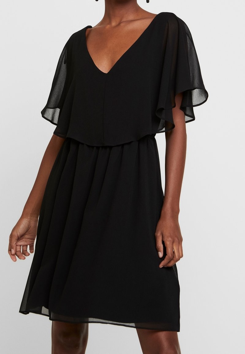 Femme portant une robe noire mi-longue avec des manches flottantes et un décolleté en V sur un fond clair uni.