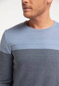 Homme portant un pull en tricot texturé bleu et gris, montré des épaules au bas du visage contre un fond blanc uni.