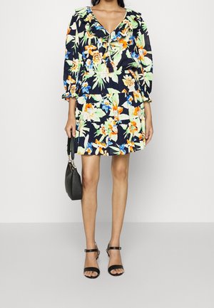 Geblümtes Kleid mit marineblauem Hintergrund, mit gerafftem Ausschnitt, langen Ärmeln und gestuftem Rock. Kombiniert mit schwarzen Riemchen-Absätzen und einer Tasche.