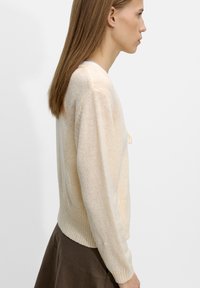 Jasnobeżowy sweter dzianinowy z prążkowanym dołem, w luźnym kroju i długimi rękawami. Tekstura sprawia wrażenie miękkiej i lekko prześwitującej.
