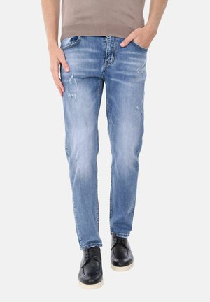 Uomo che indossa jeans azzurri slavati e scarpe nere con suole bianche, in piedi con una mano in tasca.