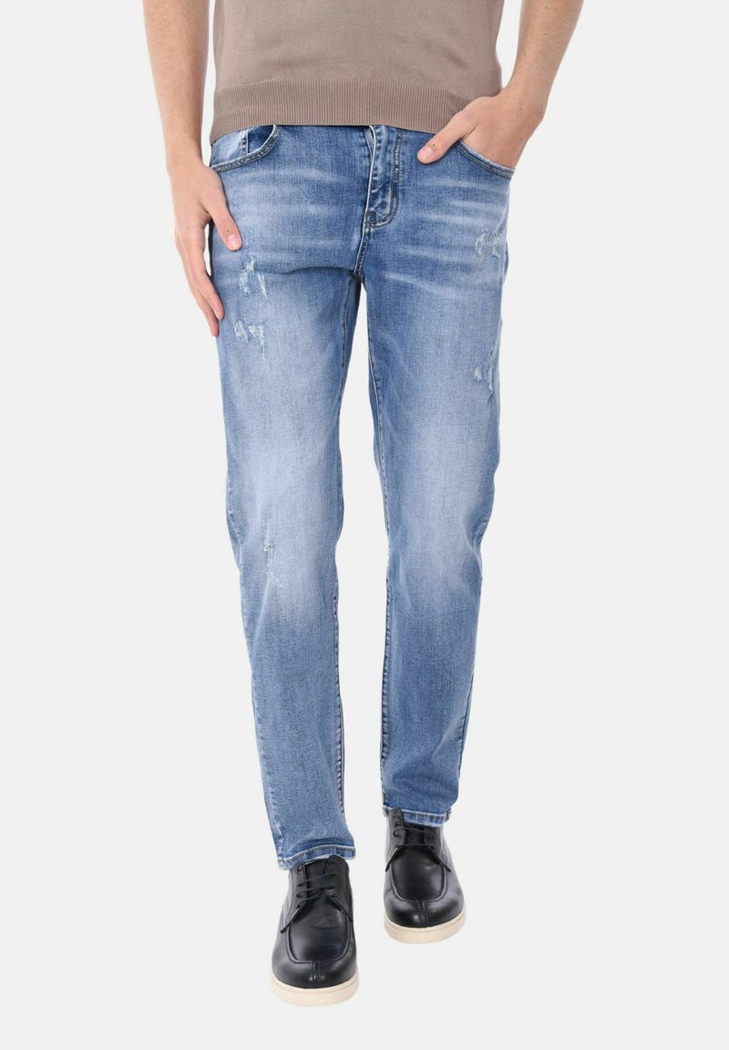 Uomo che indossa jeans azzurri slavati e scarpe nere con suole bianche, in piedi con una mano in tasca.